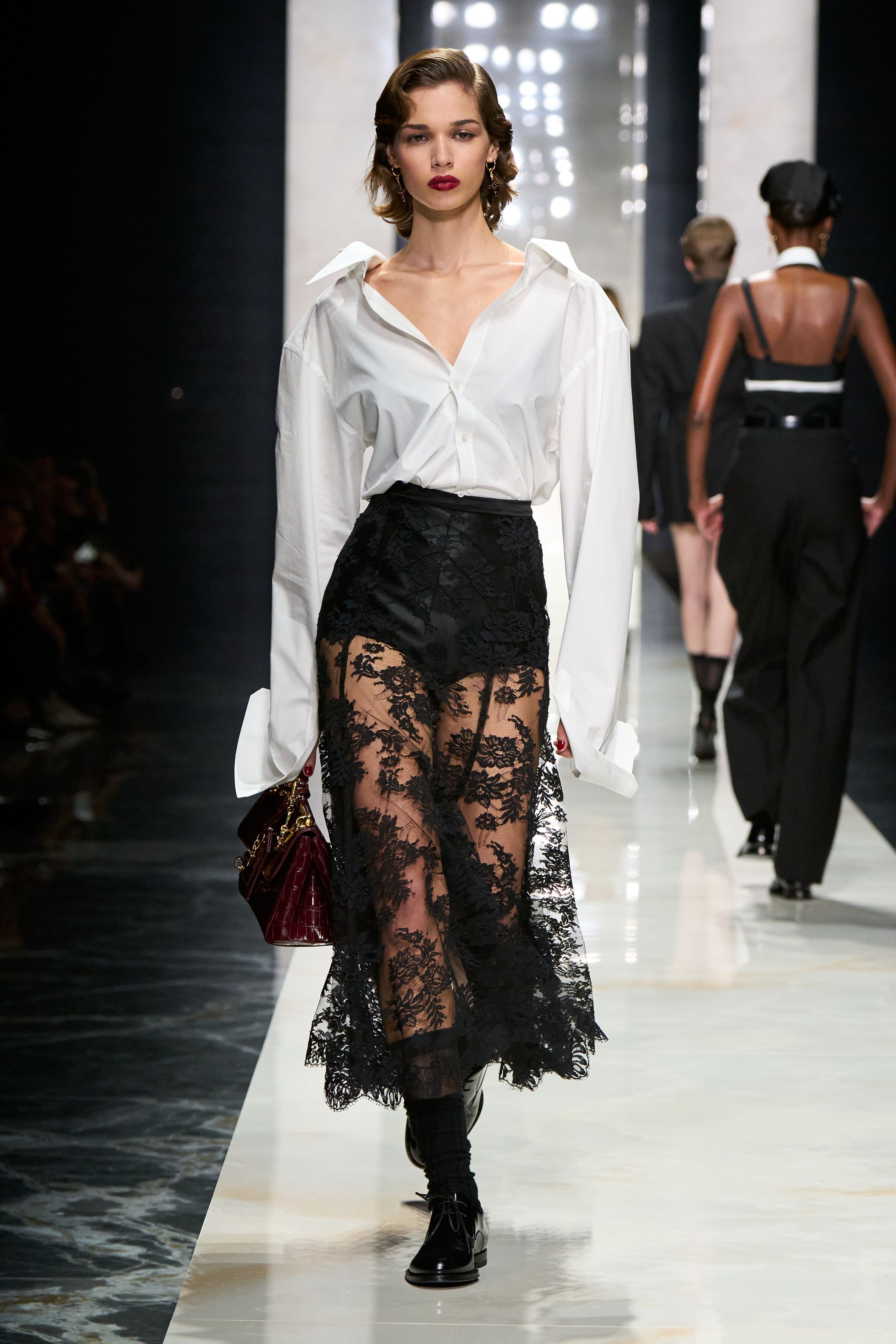 Dolce & Gabbana在本季回归品牌标志性的性感意大利美学，通过黑色蕾丝、修身西装与透视礼服塑造极具戏剧张力的女性形象。系列以黑色为核心色调，结合紧身胸衣结构、透明蕾丝裙与宽肩西装，形成刚柔并济的视觉语言。皮草外套与复古帽饰则增添奢华氛围，使整体造型在性感与优雅之间保持平衡，呈现出典型的意大利电影般的华丽气质-顽石频道趋势研究